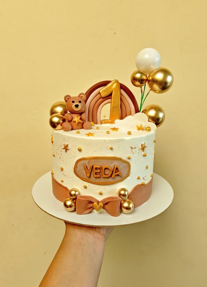 First-birthday cake in caramel tones with a teddy, rainbow and gold balloons